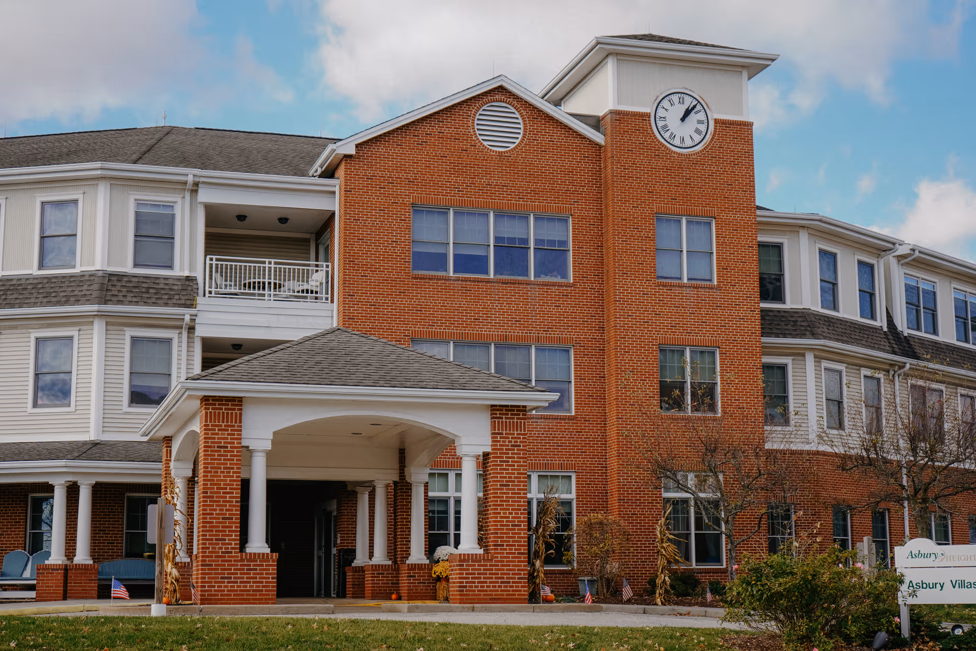 Exterior view of a multi-story senior living facility building with a brick facade and white trim. The building features a covered entrance with columns, multiple windows, and a clock on the upper right side. There is a sign in front of the building that reads Asbury Heights and Asbury Village. The sky is partly cloudy.