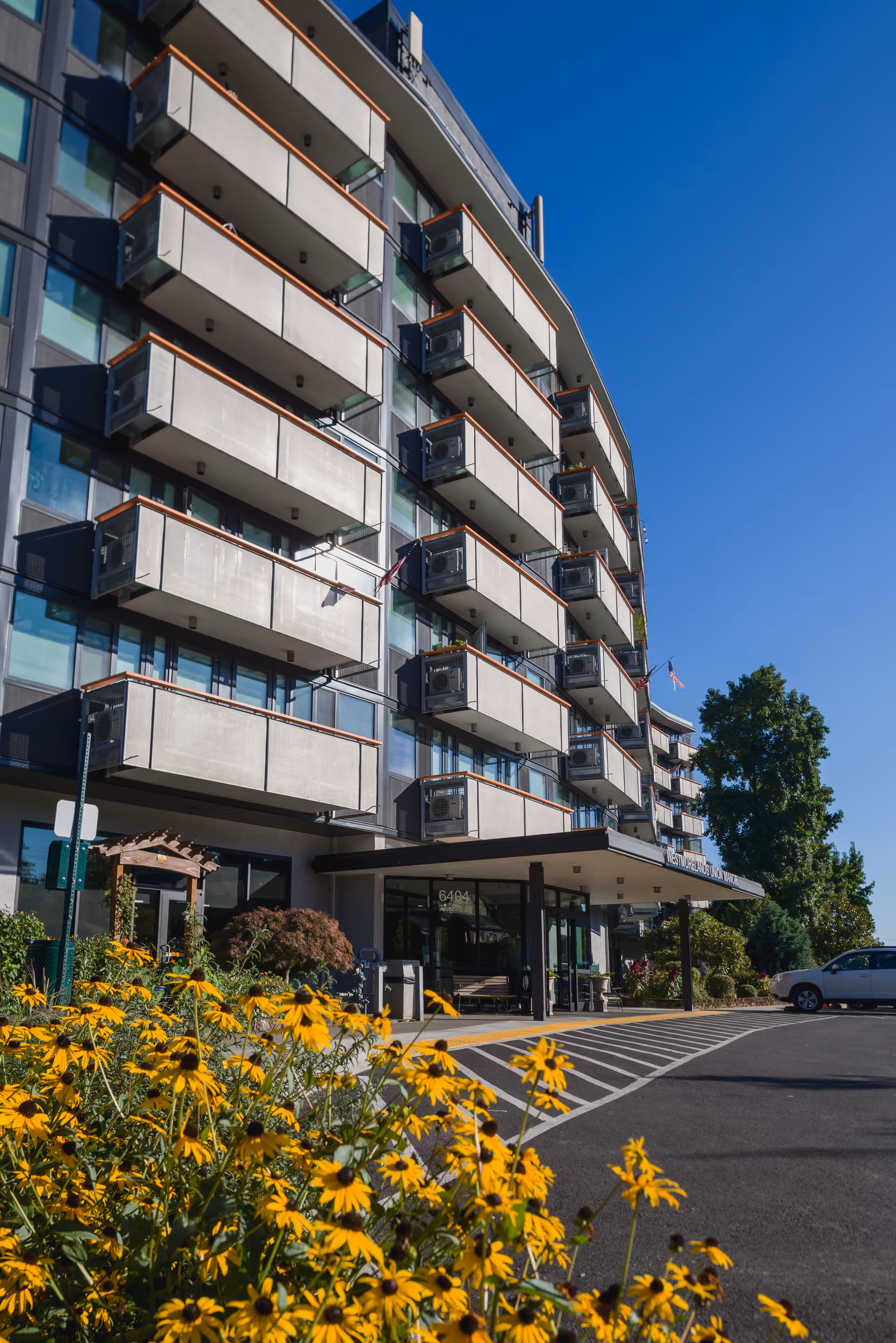Exterior view of a multi-story senior living facility with balconies on each floor, a covered entrance, and a parking area. Yellow flowers and greenery are visible in the foreground under a clear blue sky.