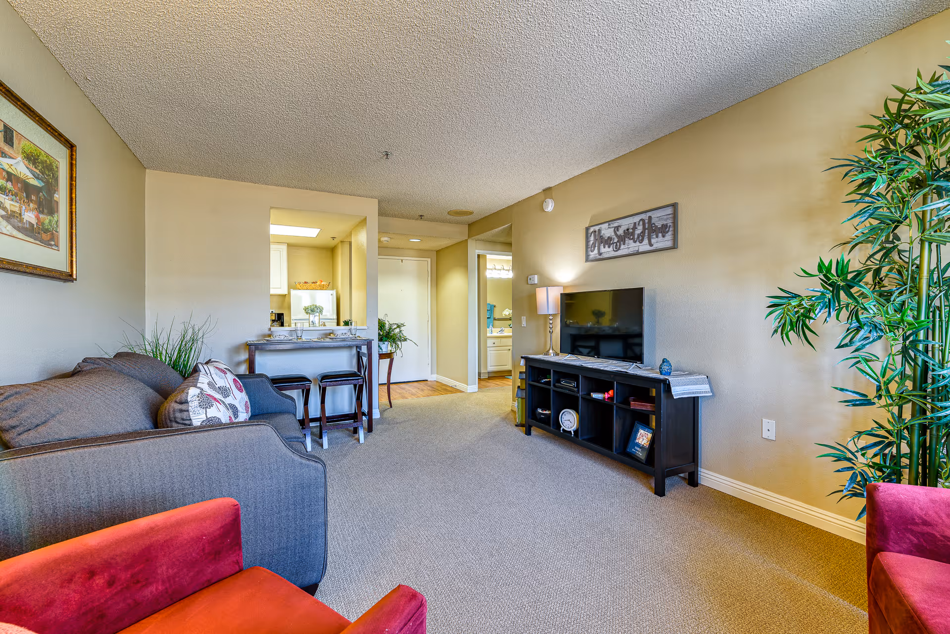 A cozy living room in a senior living facility featuring a gray sofa with patterned pillows, a red armchair, a black TV stand with a flat-screen television, a lamp, and decorative items. There is a small dining area with two stools and a counter near the kitchen entrance. The walls are painted beige, and there is a large green plant in the corner. A framed picture hangs on one wall, and a sign above the TV reads 'Home Sweet Home.'