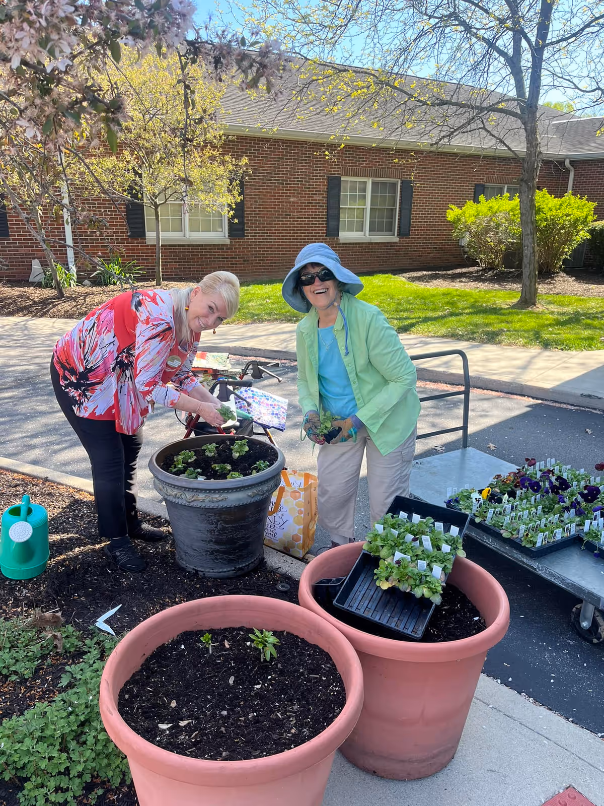 Two women are gardening outdoors in front of a brick building. One woman is wearing a red and white patterned top and black pants, bending over to plant in a large black pot. The other woman is wearing a light green jacket, blue shirt, beige pants, a blue sun hat, and sunglasses, holding plants and smiling. There are large empty and partially filled flower pots, a green watering can, and trays of plants on a cart nearby. Trees and shrubs are visible in the background under a clear sky.