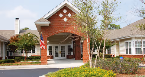 Front exterior view of a senior living facility named Brookdale Wilmington featuring a brick entrance with an American flag, surrounded by trees, shrubs, and a well-maintained garden area.