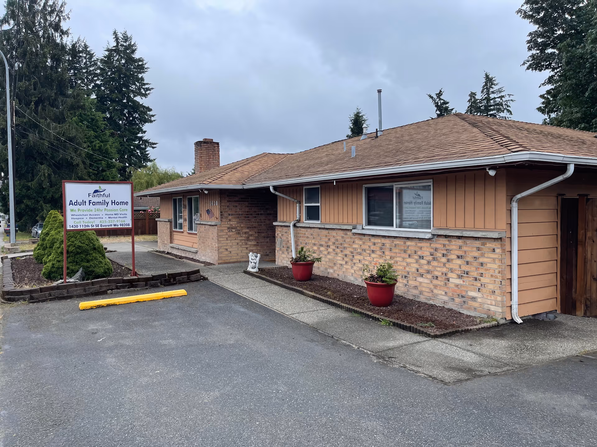 Exterior view of a single-story brick and wood-paneled building with a sloped roof, two red flower pots with plants near the entrance, and a sign that reads 'Faithful Adult Family Home' with contact information. The sky is overcast and there are trees in the background.