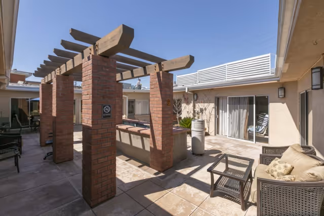 Outdoor patio area at Brookfield Healthcare Center featuring brick columns with wooden beams overhead, cushioned wicker chairs, a glass-top table, and sliding glass doors leading inside. The area is well-lit with natural sunlight and has a no smoking sign on one of the brick columns.