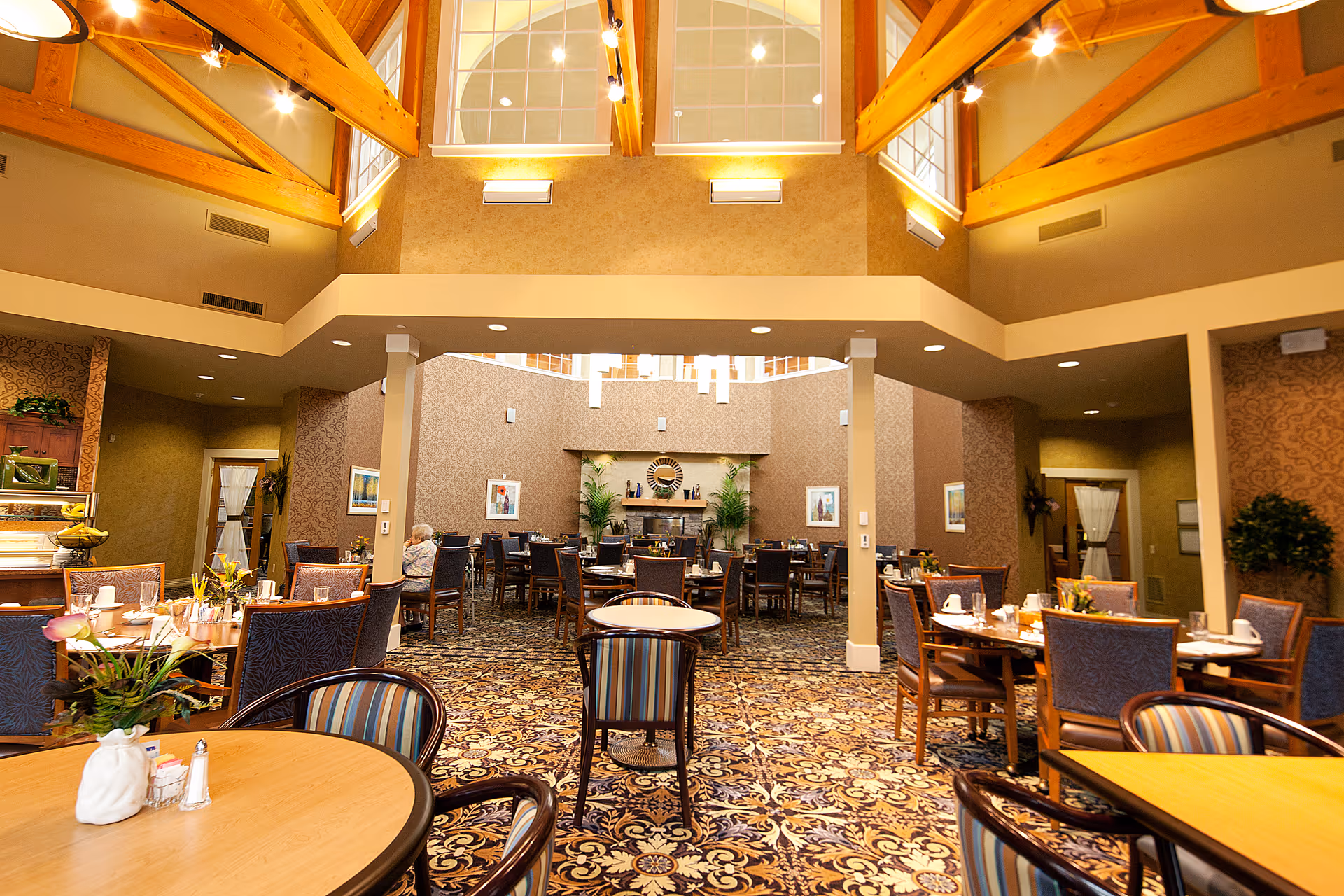 A spacious and well-lit dining room with high ceilings featuring wooden beams and large windows. The room is furnished with multiple round and square tables surrounded by chairs, set with cups, plates, and small flower arrangements. The carpet has an intricate floral pattern, and the walls are decorated with framed artwork and plants. Warm lighting creates a cozy atmosphere.