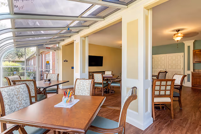 A bright and inviting indoor seating area with wooden tables and cushioned chairs featuring floral upholstery. The space has large windows and a glass ceiling allowing natural light to fill the room. In the background, there is a wall-mounted television and additional seating areas with similar chairs and tables. The flooring is wooden, and the walls are painted in soft neutral tones.