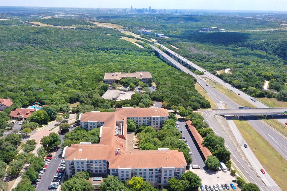Aerial view of Brookdale Gaines Ranch facility surrounded by dense greenery and trees, with a highway and overpass visible to the right and a city skyline in the distant background.
