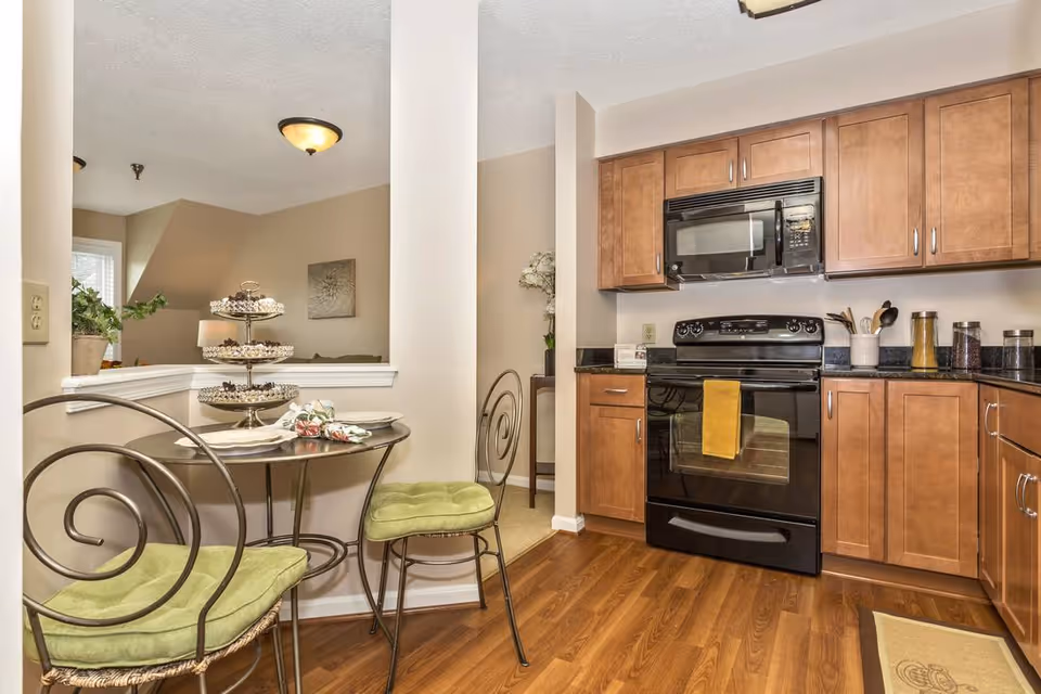 A cozy kitchen area with wooden cabinets, a black stove and microwave, and a small round table with two metal chairs that have green cushions. The table is set with plates, napkins, and a tiered tray of cupcakes. The floor is wood, and there is a partial wall separating the kitchen from an adjacent room.