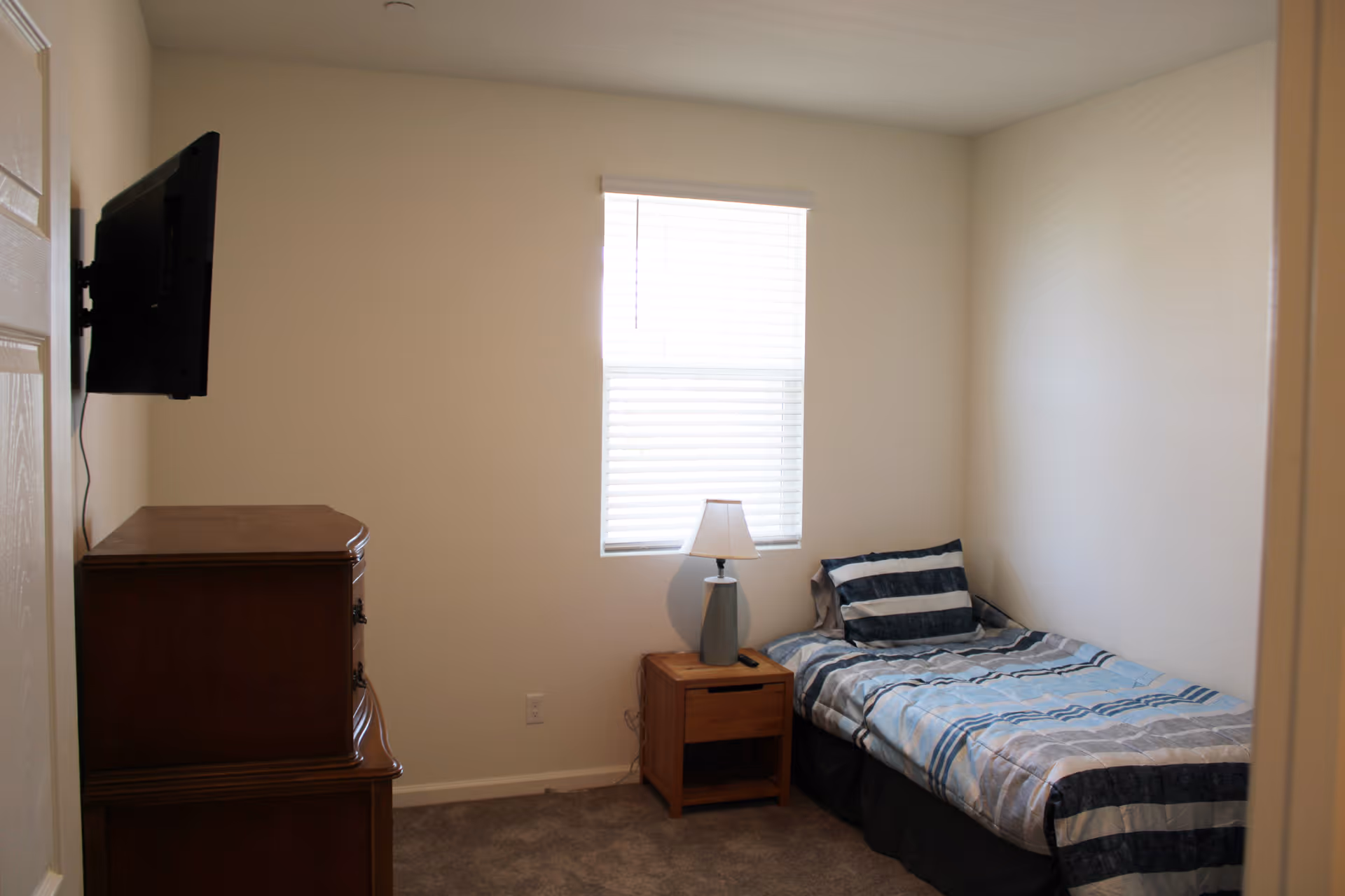 A small bedroom with a single bed covered in a blue and gray striped comforter and matching pillow. Next to the bed is a wooden nightstand with a lamp and a remote control. A wooden dresser is positioned against the opposite wall, and a flat-screen TV is mounted above it. A window with white blinds is centered on the wall behind the bed.