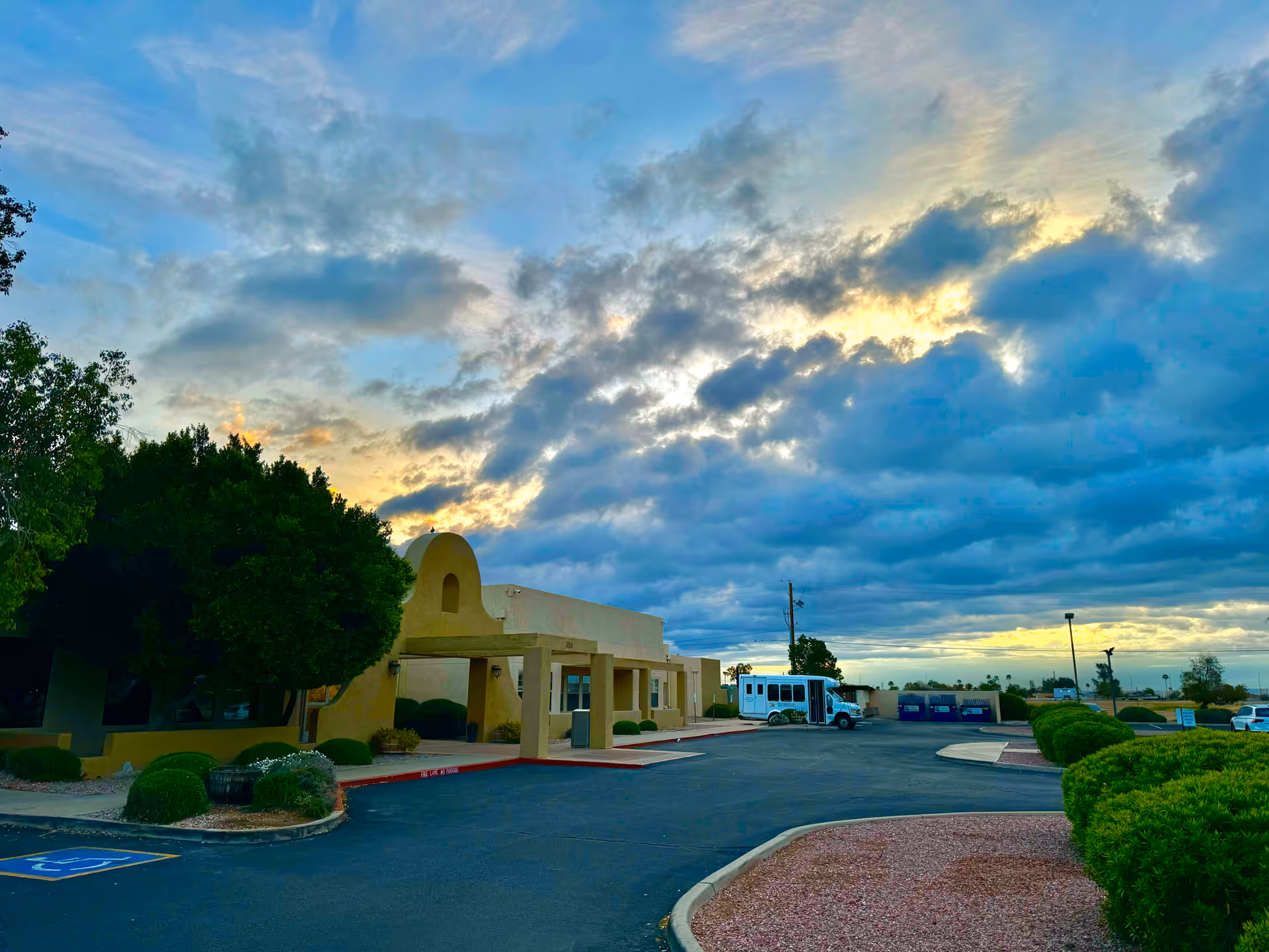 Exterior view of Estrella Health and Rehab Center building during sunset with a partly cloudy sky, a parking lot with a bus, and landscaped bushes and trees.