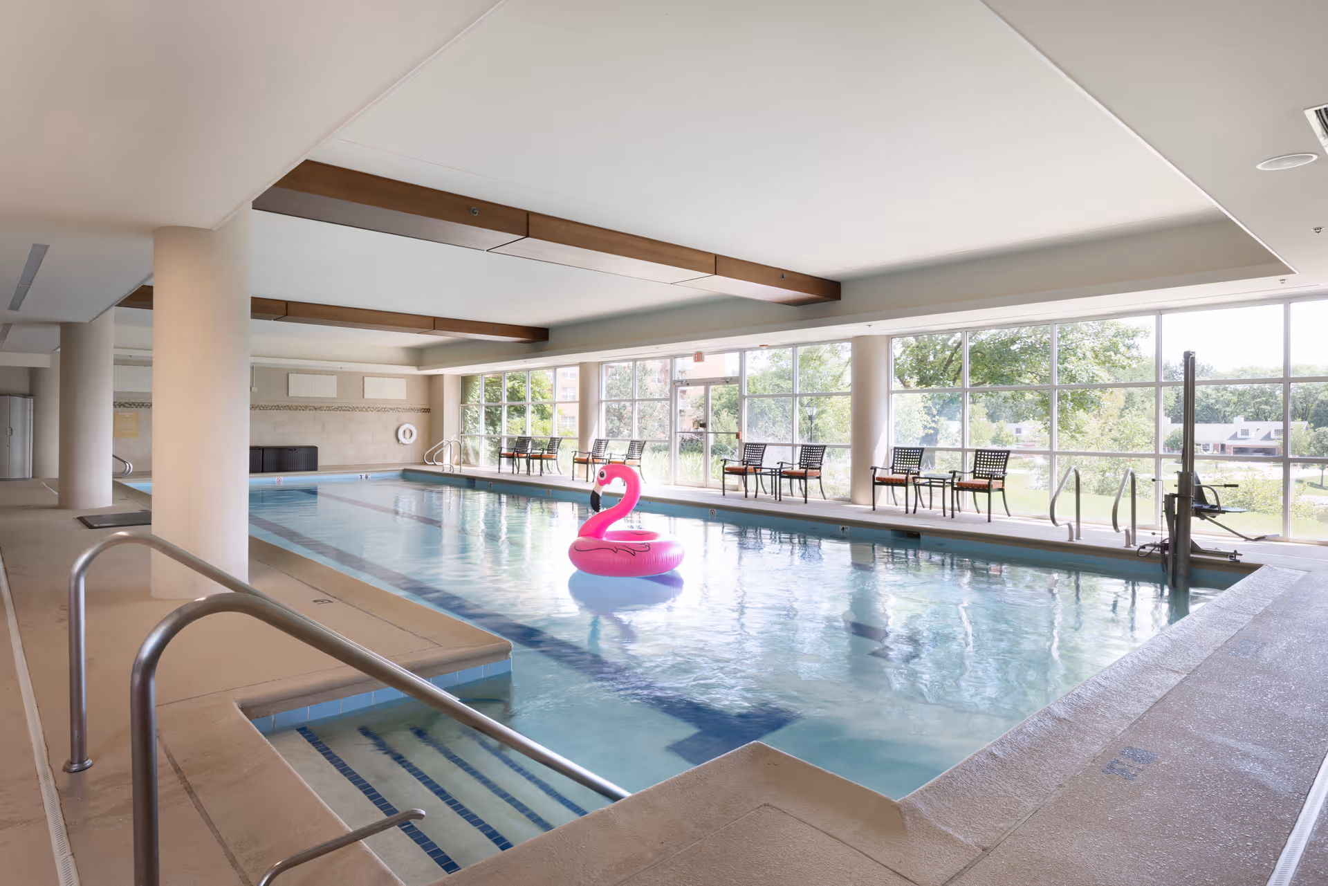 Indoor swimming pool with a pink flamingo float in the water. The pool area has large windows letting in natural light, several chairs and tables along the side, and beige tiled flooring with metal handrails leading into the pool.