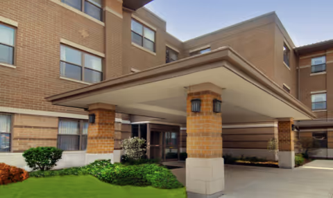 Exterior view of a multi-story brick building with a covered entrance supported by brick columns. There are windows on each floor and some landscaping with green bushes and grass near the entrance.