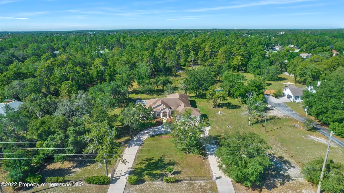 Aerial view of a single-story assisted living facility building surrounded by lush green trees and open grassy areas. The building has a curved driveway leading to the entrance, and there are other smaller structures visible nearby. The area is peaceful and spacious with a dense forest in the background under a clear blue sky.