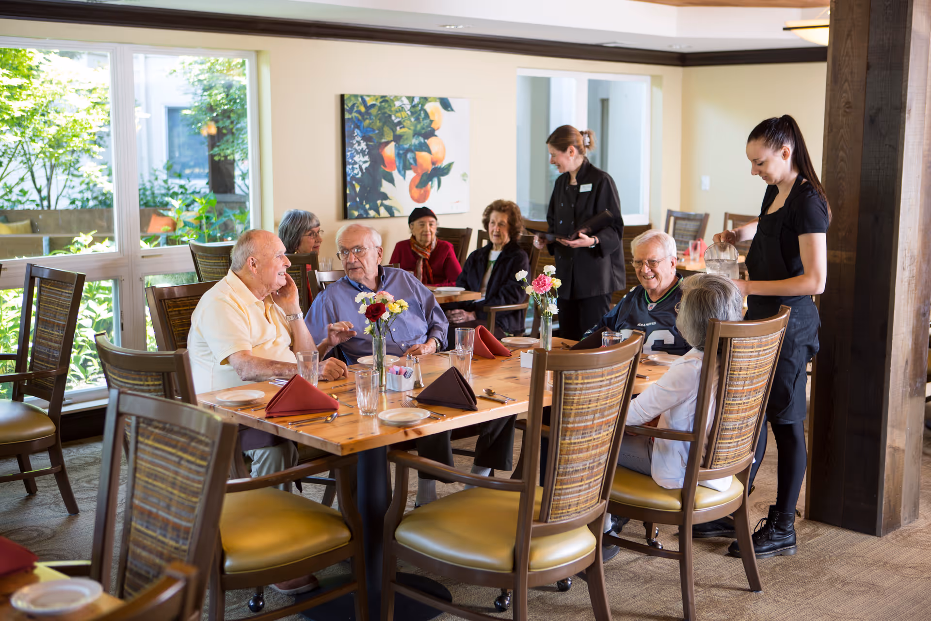 A group of elderly people sitting around a dining table in a well-lit room with large windows and greenery outside. Two staff members are attending to them, one pouring water and the other holding a tray. The table is set with plates, glasses, and napkins, and there are flower vases as centerpieces.