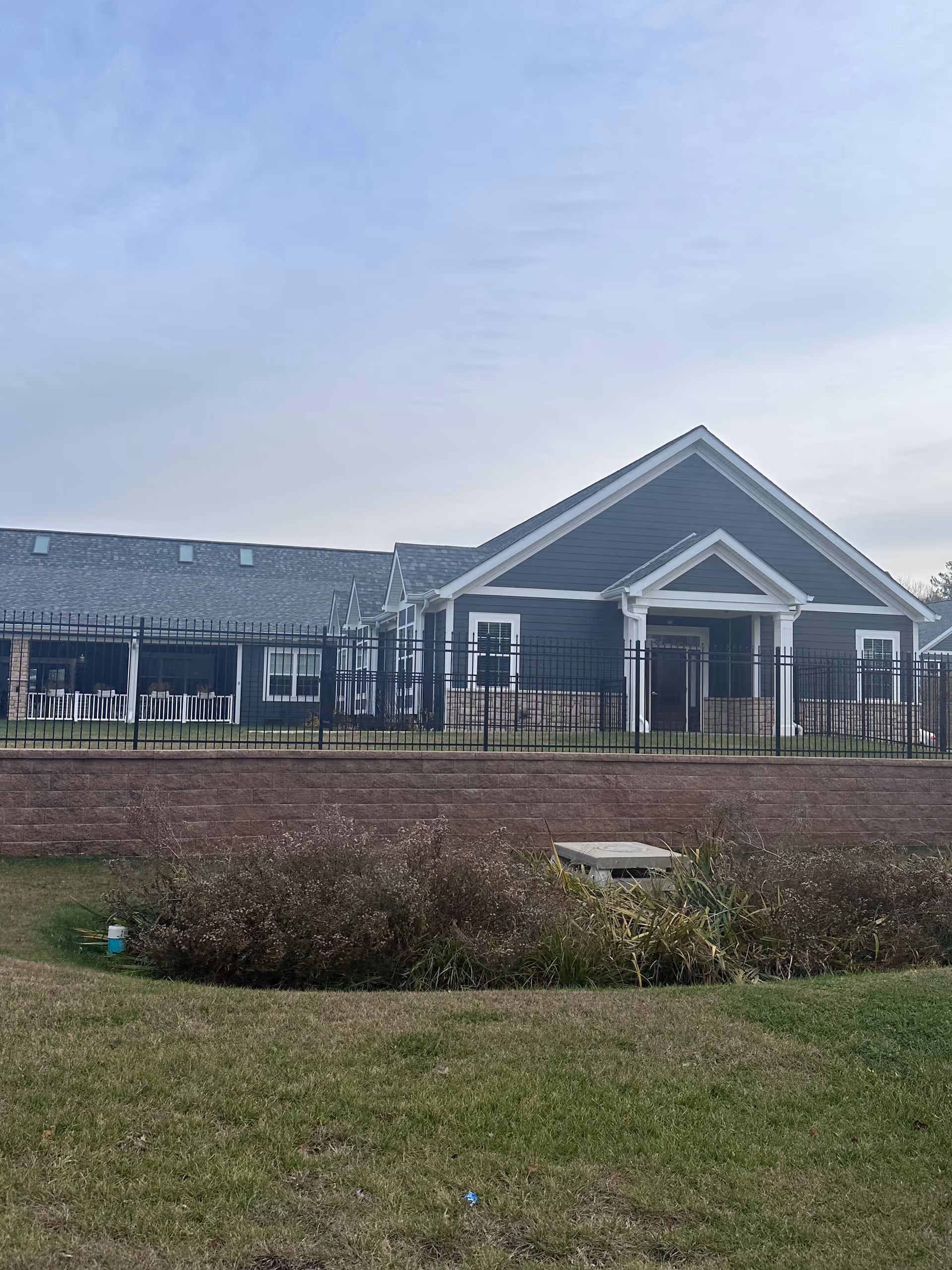 Exterior view of a single-story building with dark blue siding and white trim, featuring a gabled roof and a covered entrance. The building is surrounded by a black metal fence and a retaining wall with landscaping in front. The sky is partly cloudy.