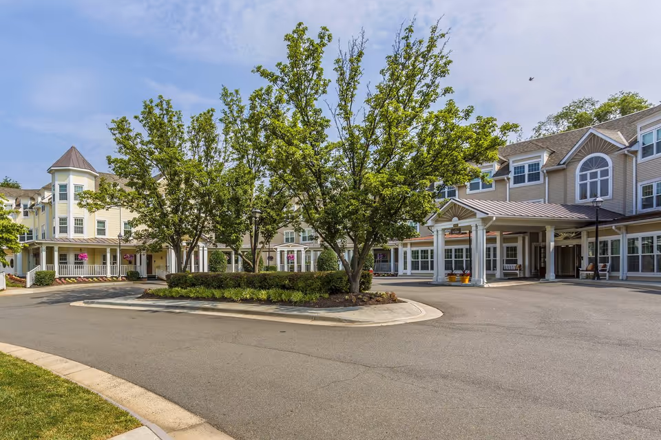 Exterior view of a senior living facility with a circular driveway, landscaped greenery, and trees in front of a multi-story building with large windows and a covered entrance.
