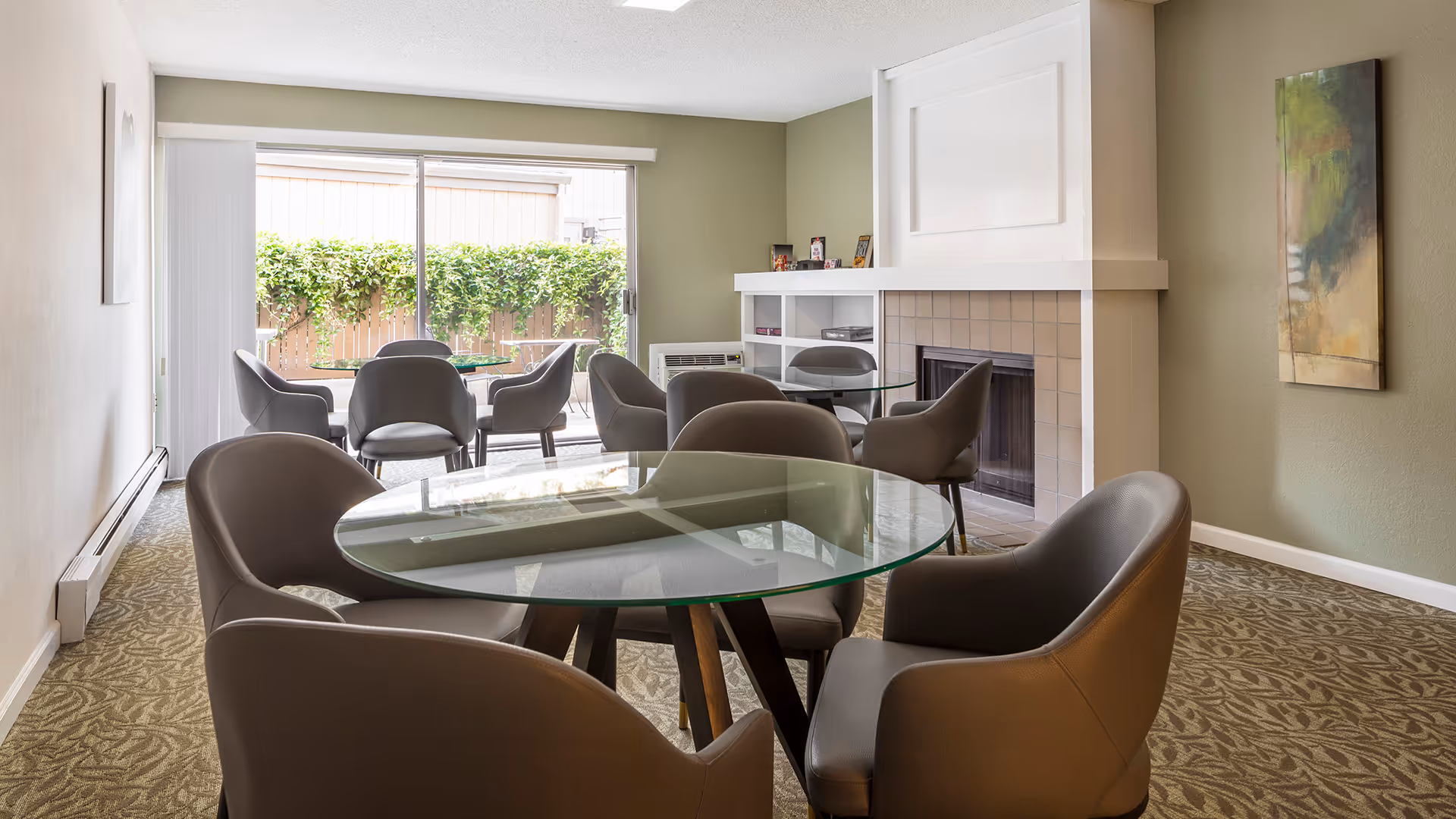 A bright and modern common area with multiple round glass tables surrounded by gray cushioned chairs. The room features a fireplace with a white mantel, a large window with vertical blinds letting in natural light, and a green wall with a colorful abstract painting. Outside the window, there is a wooden fence with green vines.