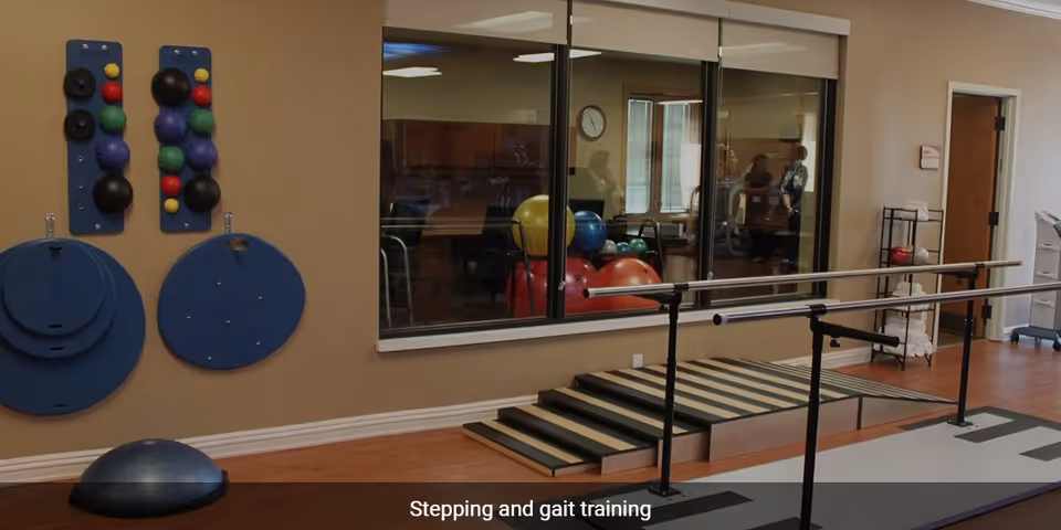 Physical therapy room with parallel bars for gait training, a small set of stairs with a ramp, balance training equipment mounted on the wall, and exercise balls visible through a large window into an adjacent room.