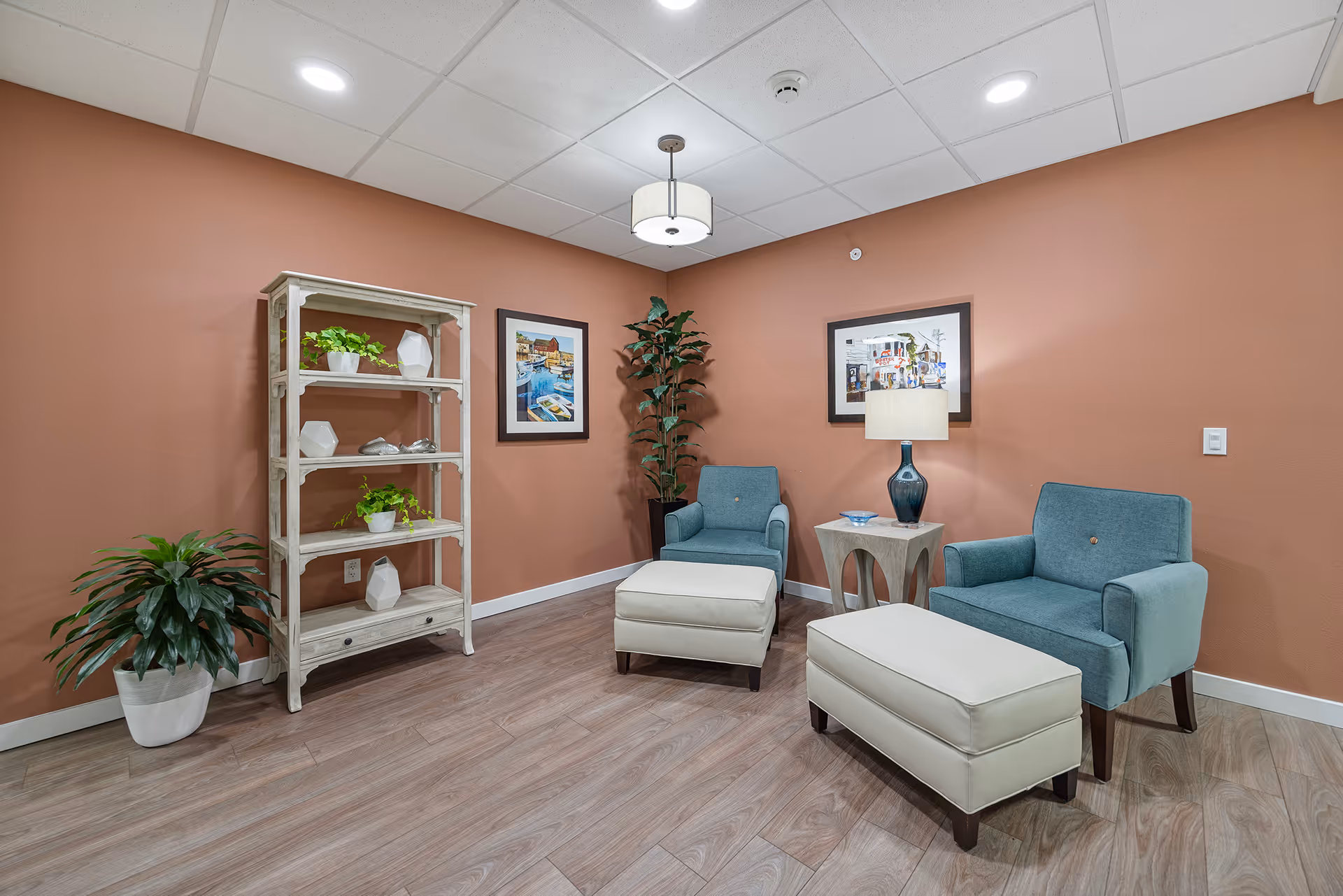 A cozy seating area with two blue armchairs and two white ottomans on a wood floor. There is a small side table with a blue lamp between the chairs. The walls are painted a warm terracotta color and decorated with two framed pictures. A white shelving unit with decorative items and two potted plants are also visible. The ceiling has recessed lighting and a hanging light fixture.