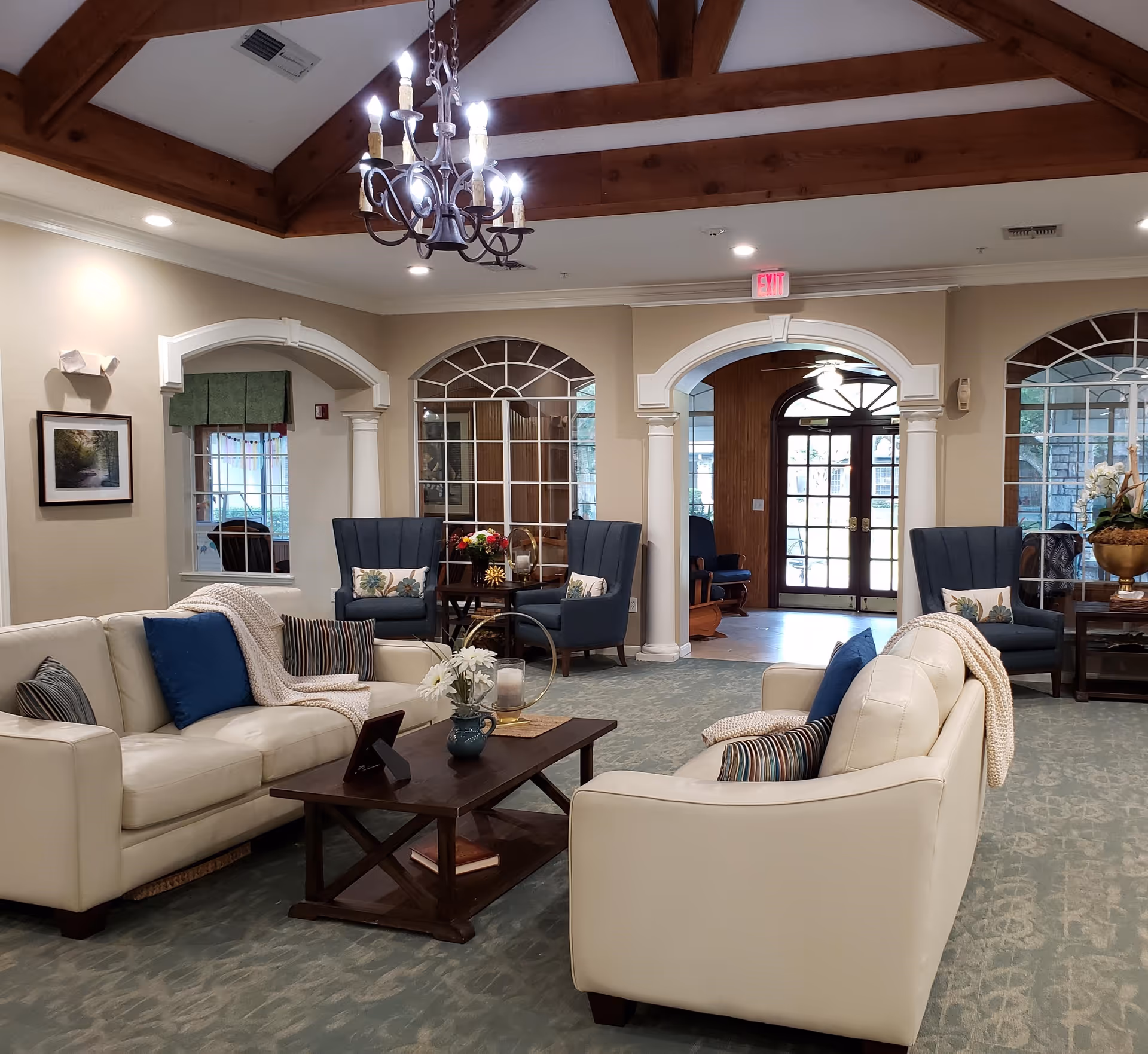 A cozy living room area in a senior living facility with two white leather sofas facing each other, a wooden coffee table with decorative items, and several blue upholstered armchairs. The room features a high ceiling with exposed wooden beams and a chandelier, large windows with decorative arches, and a glass door leading outside.