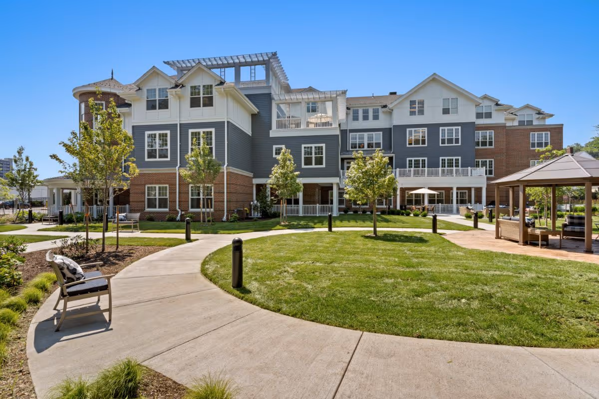 Outdoor view of a senior living facility with a curved concrete walkway, green lawn, young trees, benches, and a gazebo with seating. The building has multiple stories with a combination of brick and gray siding, white trim, and several windows under a clear blue sky.