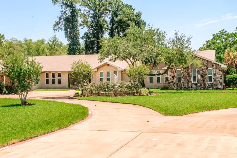 Exterior view of a single-story assisted living facility building with a stone and brick facade, surrounded by green grass, trees, and shrubs under a clear blue sky.