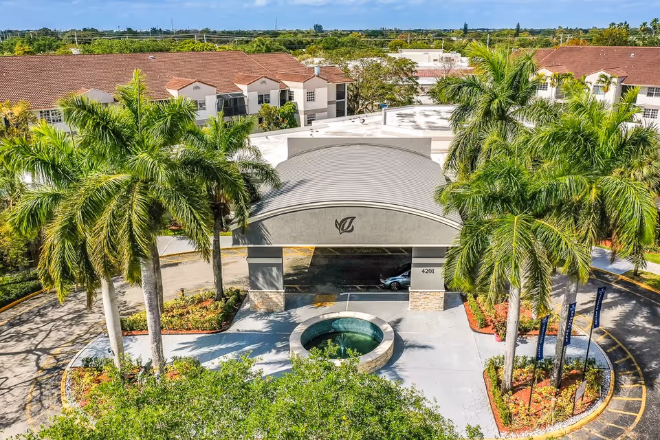 Aerial view of the entrance to Springtree Senior Living facility featuring a covered driveway with a circular fountain in front, surrounded by palm trees and landscaped greenery. The building has a beige exterior with a red-tiled roof and multiple windows.