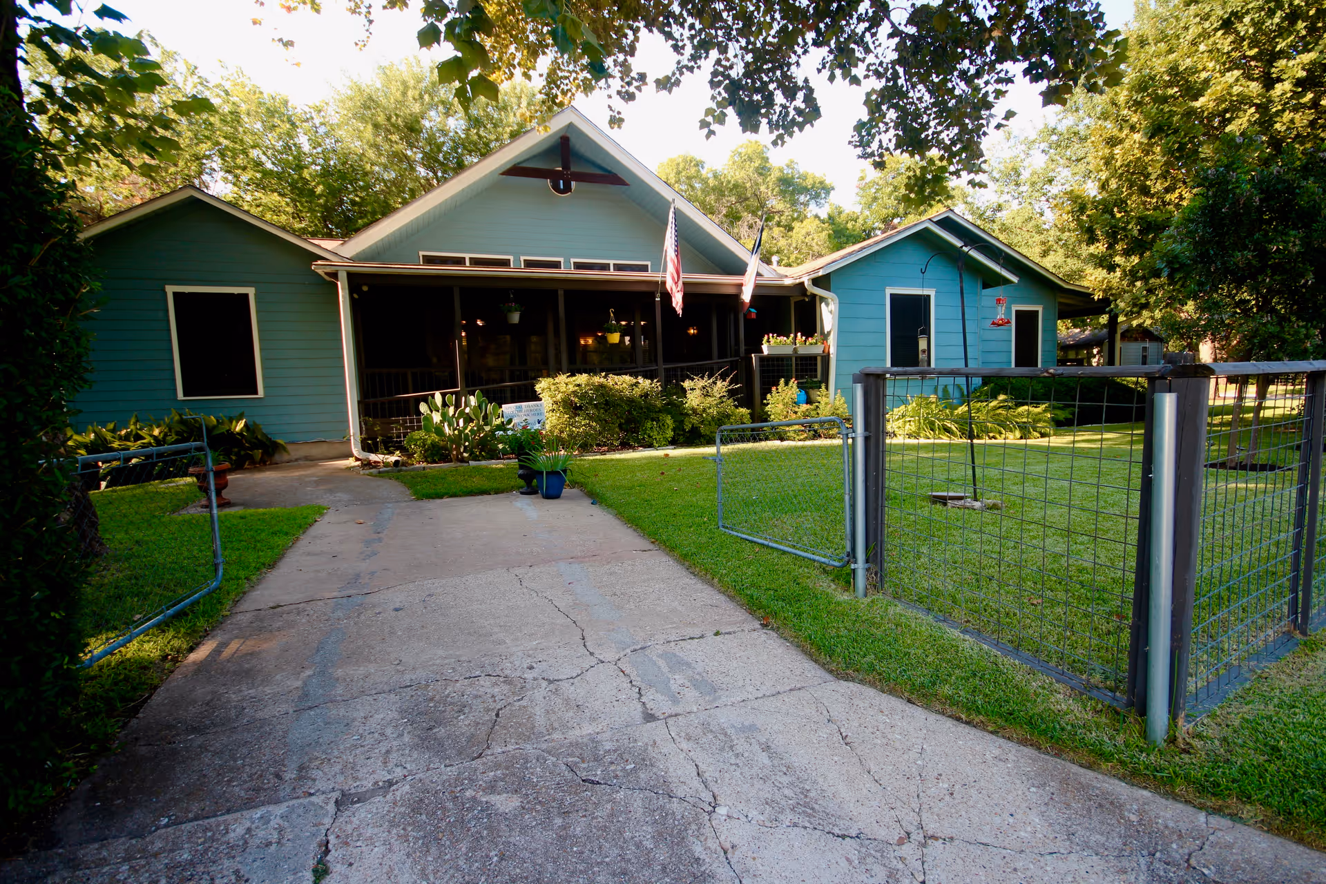 Front exterior view of a single-story blue house with a driveway leading up to a fenced yard. The house has a covered porch with hanging plants and two flags displayed. The yard is well-maintained with green grass and trees surrounding the property.