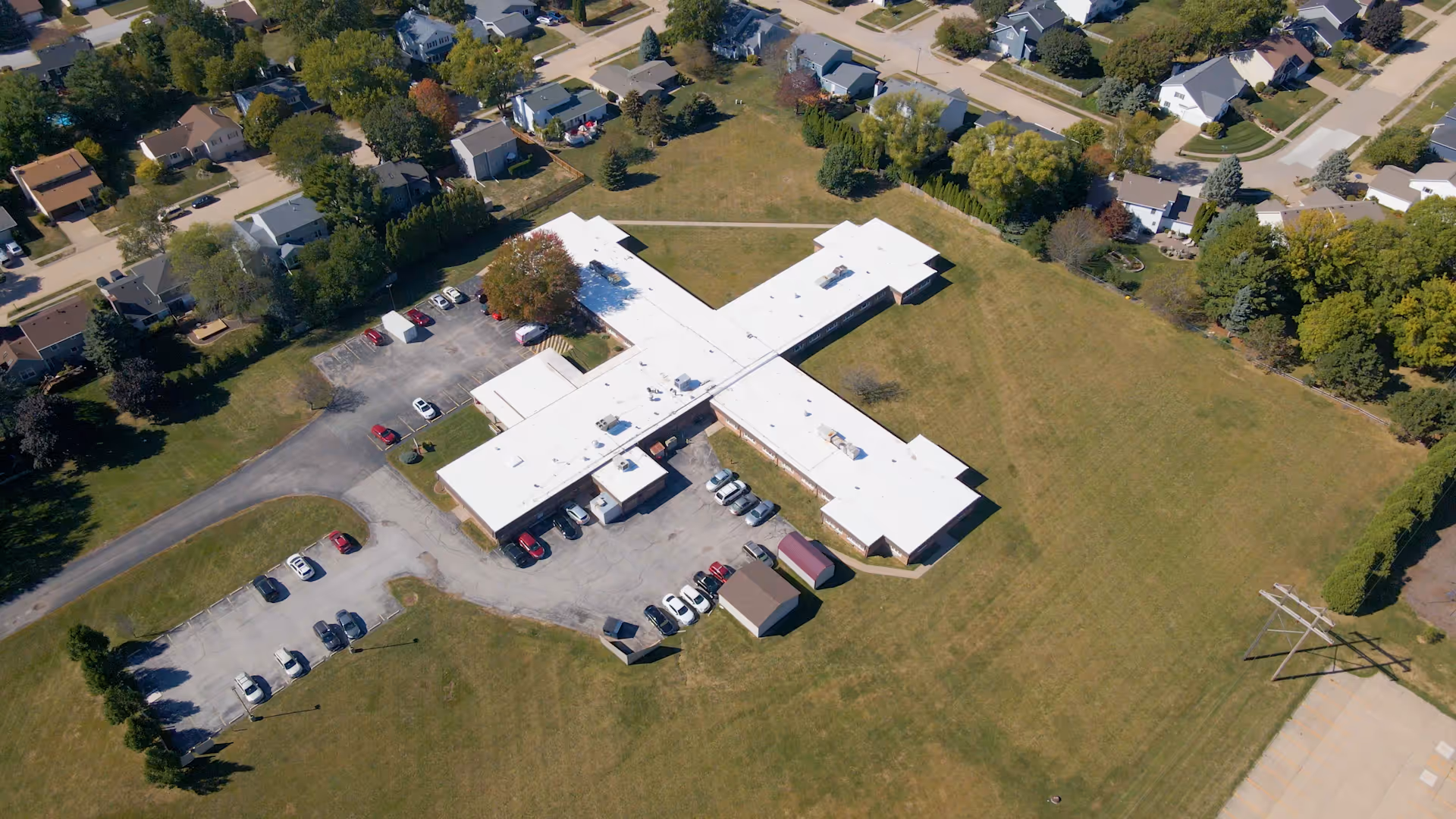 Aerial view of a nursing and rehab center building with a white roof shaped like a cross, surrounded by a large grassy area and parking lots with several cars. Residential houses and trees are visible in the background.