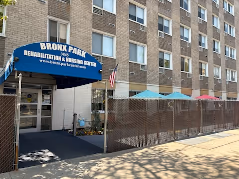 Exterior view of Bronx Park Rehabilitation & Nursing Center showing the entrance with a blue awning displaying the facility name and website. The building is a multi-story brick structure with several windows. There is a fenced area with colorful umbrellas visible to the right of the entrance.