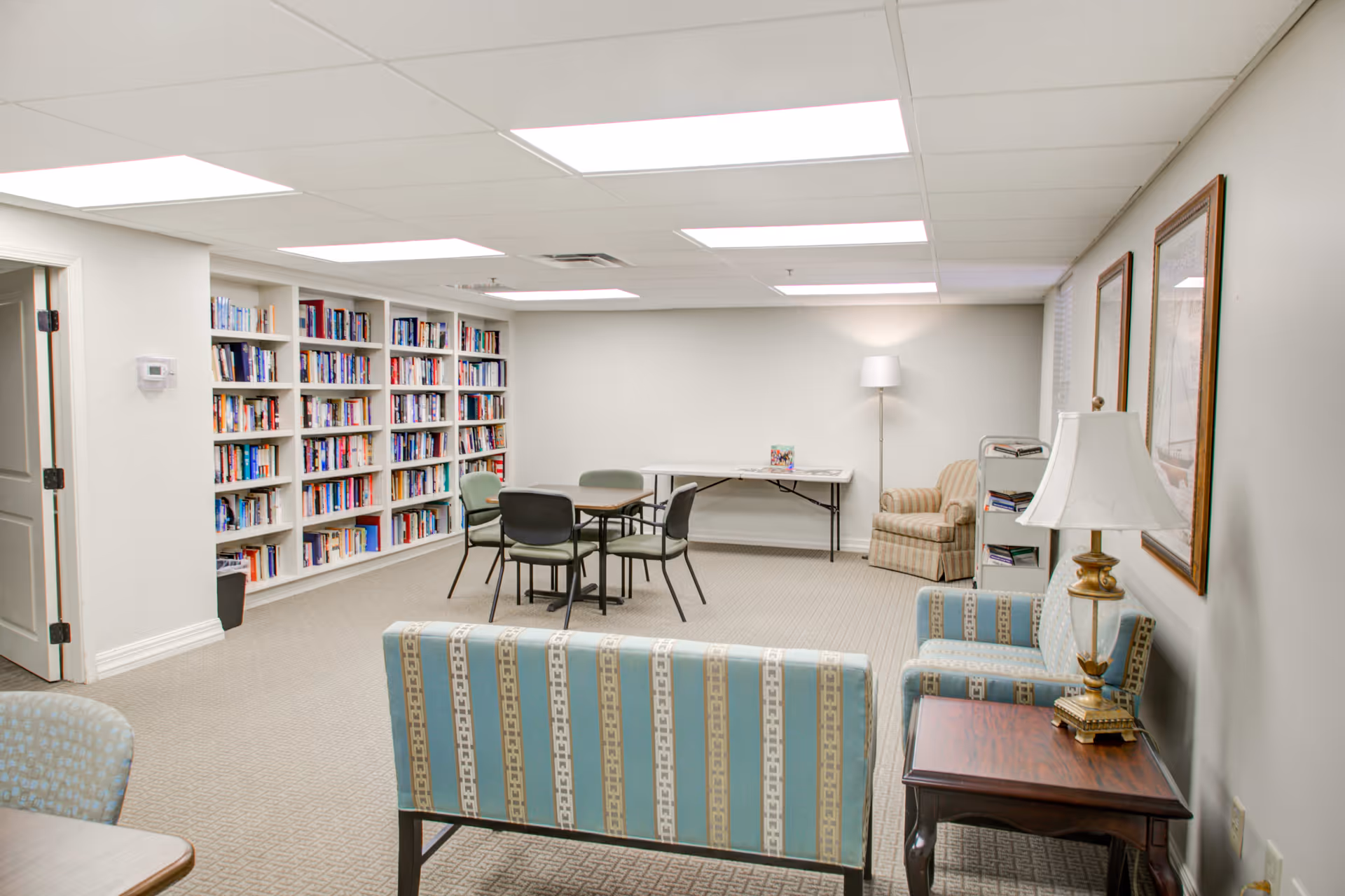 A well-lit room with bookshelves filled with books along one wall, a table with four chairs in the center, a striped armchair and a floor lamp in the corner, and two upholstered chairs with a small wooden side table and lamp in the foreground. The walls are light-colored and the ceiling has recessed lighting.