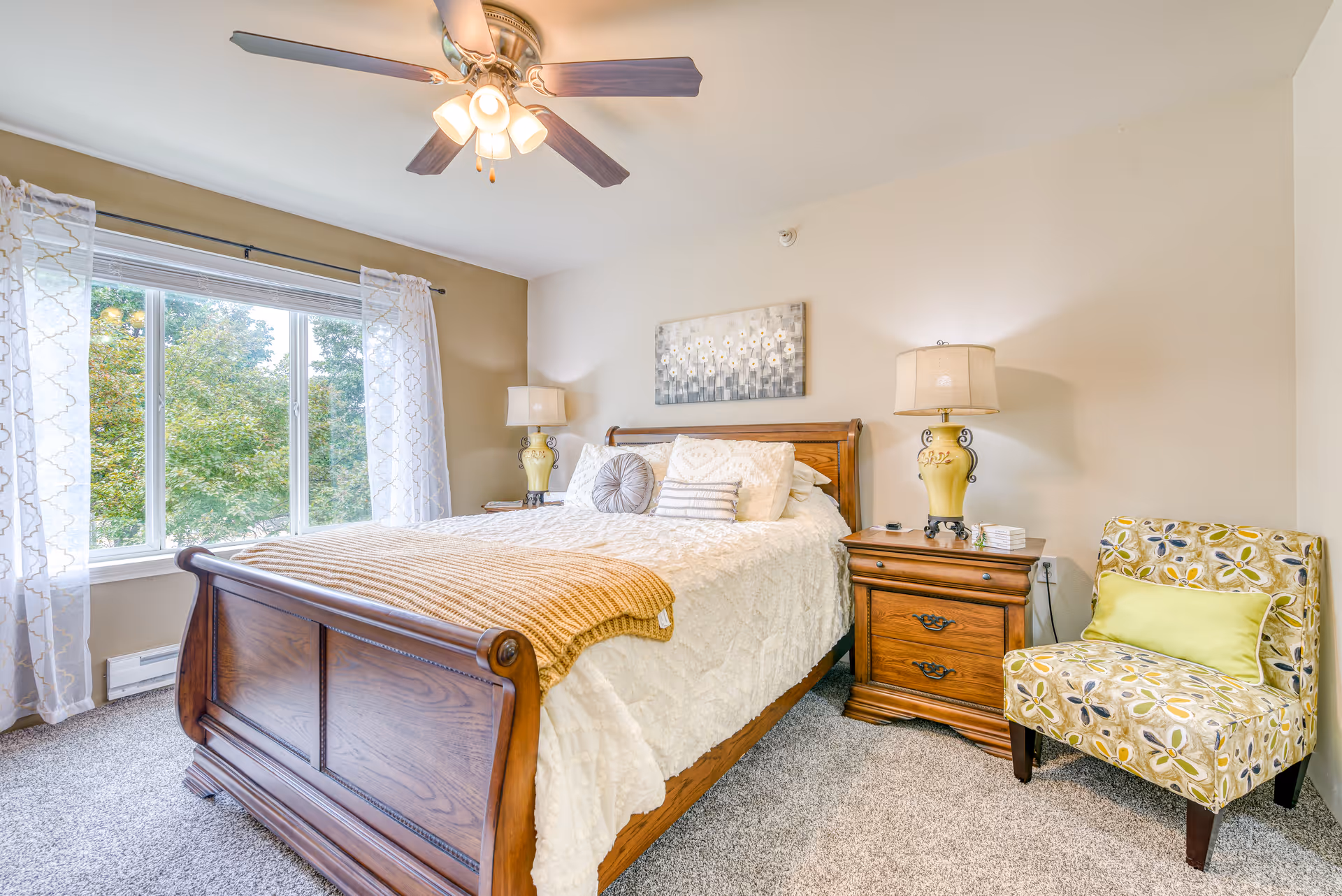 A bright and cozy bedroom featuring a wooden bed with white bedding and decorative pillows, a wooden nightstand with a lamp, a patterned armchair with a green cushion, a ceiling fan with lights, and a large window with sheer white curtains overlooking green trees.
