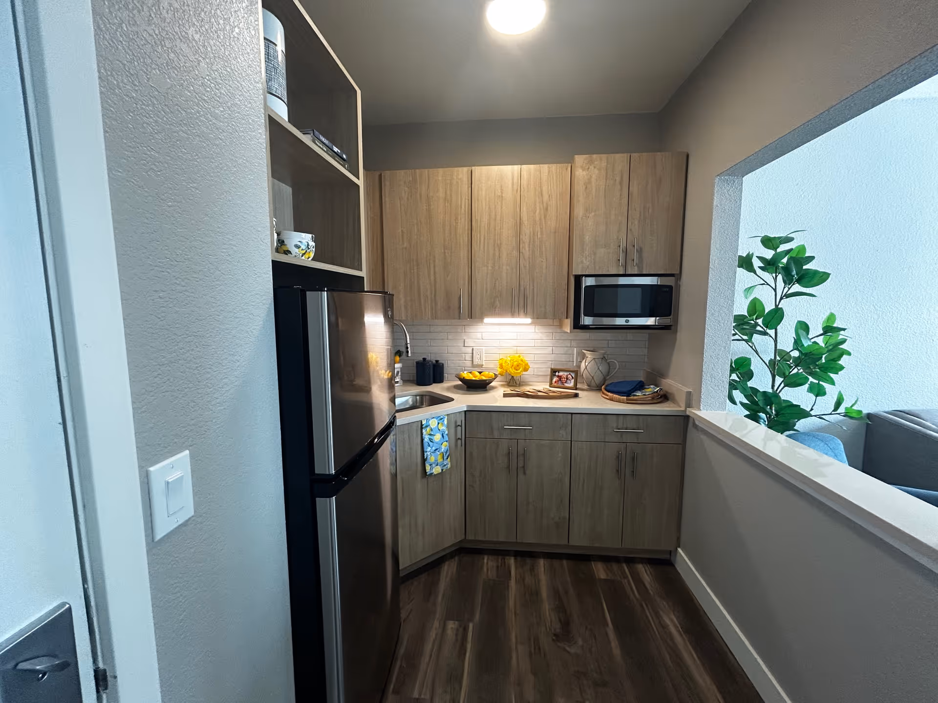 A modern kitchen with wooden cabinets, a stainless steel refrigerator, a microwave, a sink, and a countertop decorated with a bowl of lemons, a vase of yellow flowers, a framed photo, and some kitchen towels. The kitchen has dark wood flooring and a pass-through window opening to a living area with a green plant visible.
