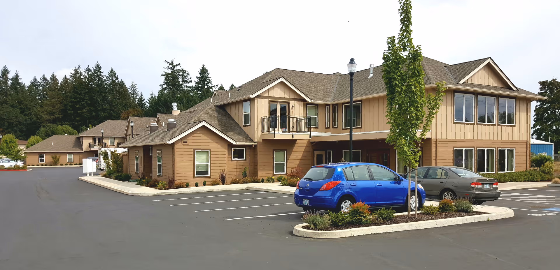 Exterior view of a two-story senior living facility building with beige and brown siding, several windows, a small balcony, and a parking lot with a few cars parked. There are trees and landscaped areas around the building.