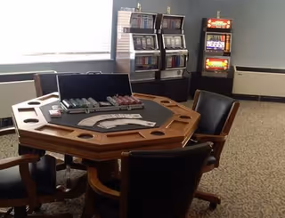 A room with a wooden octagonal poker table surrounded by four black leather chairs. The table has poker chips and playing cards on it. In the background, there are three slot machines against the wall. The room has carpeted flooring and a window with blinds partially open.
