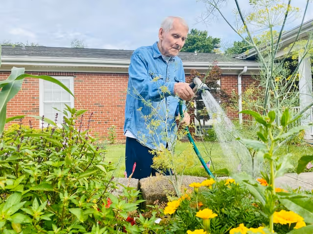 An elderly man wearing a blue denim shirt waters plants and flowers in a garden outside a brick building on a cloudy day.