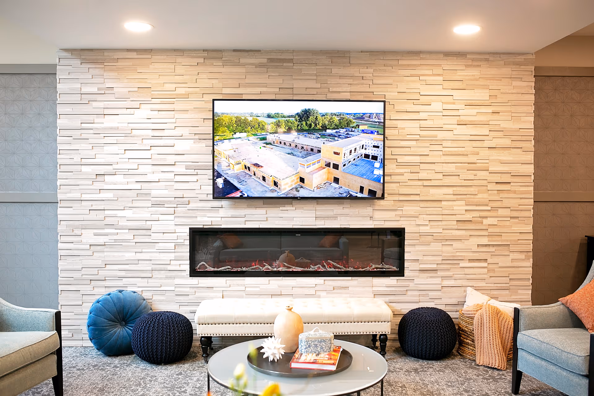 A cozy living room area with a modern electric fireplace set into a textured white stone wall. Above the fireplace is a flat-screen TV displaying an aerial view of a building complex. The room features comfortable seating including a light blue armchair, a white cushioned bench, and round knitted poufs in blue and black. A round glass coffee table with decorative items is in the foreground.