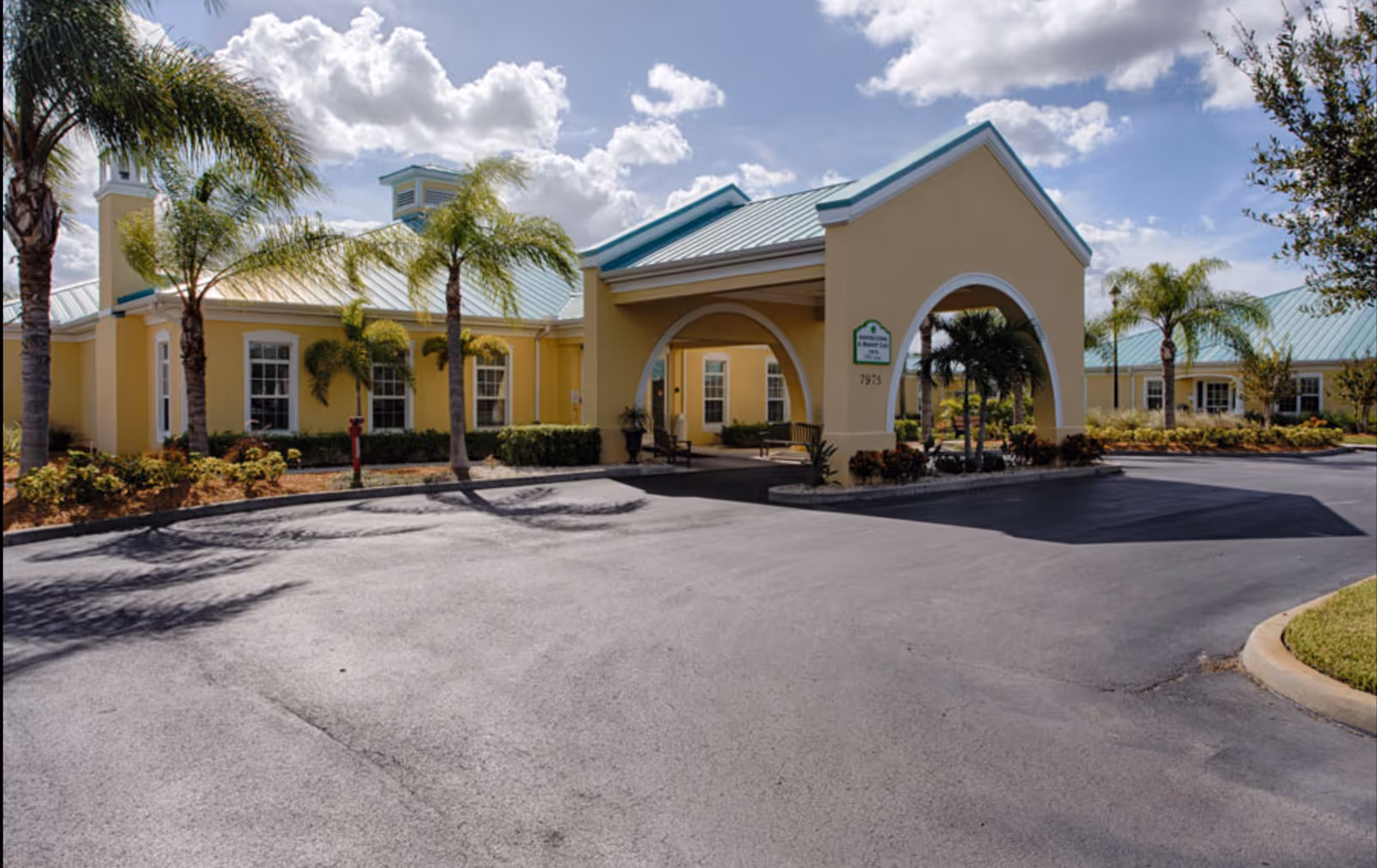 Exterior view of The Brennity at Vero Beach Senior Living building with a covered entrance, palm trees, and a clear blue sky with clouds.