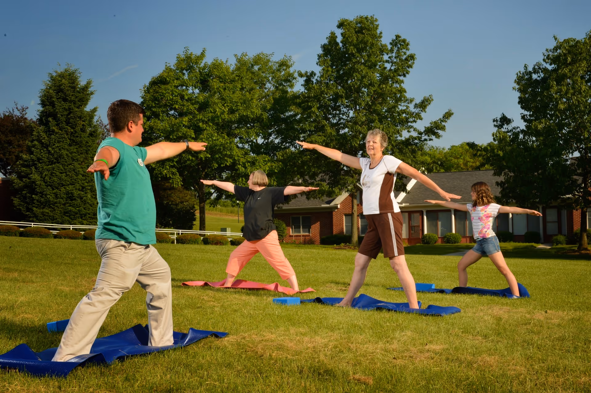 Four people practicing yoga outdoors on blue and pink mats on a grassy area with trees and a building in the background, under a clear blue sky.