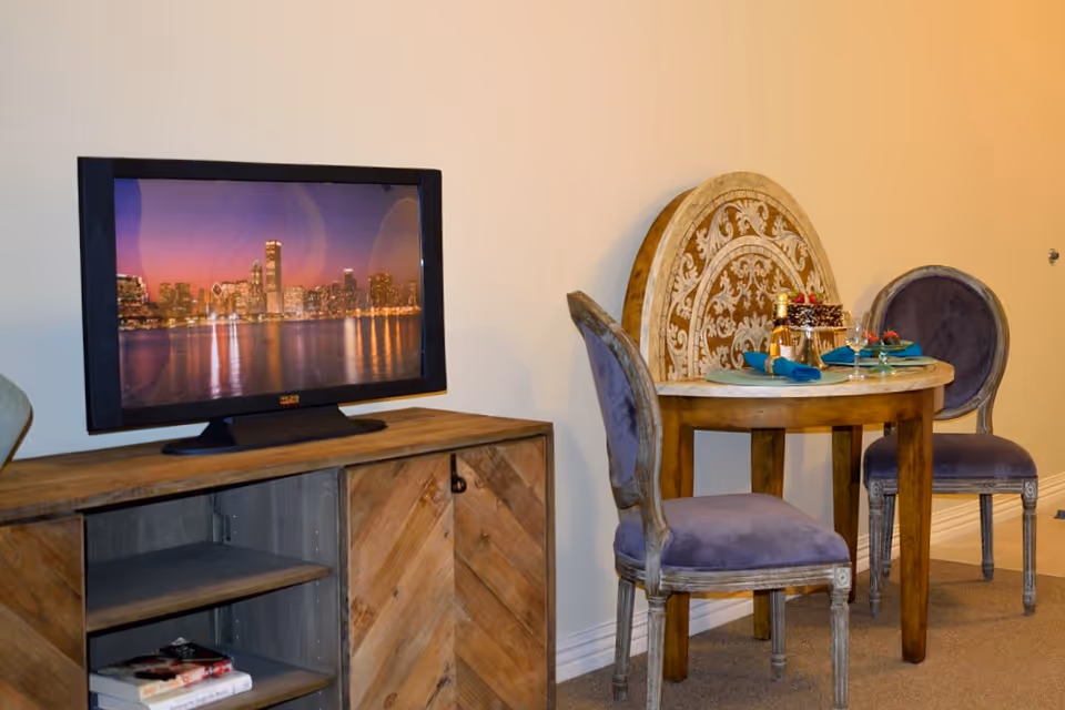 A small round wooden dining table with an ornate carved back panel, set with two place settings including plates, glasses, and napkins. Two upholstered chairs with purple cushions are placed around the table. To the left, there is a wooden cabinet with a flat-screen TV on top displaying a city skyline at dusk.