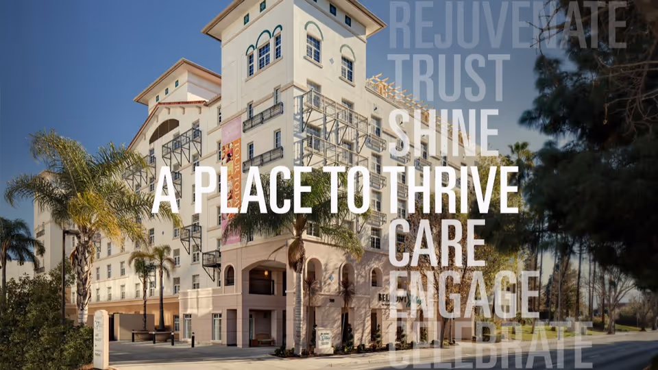 Exterior view of Belmont Village Senior Living San Jose, a multi-story building with balconies and palm trees in front under a clear blue sky.