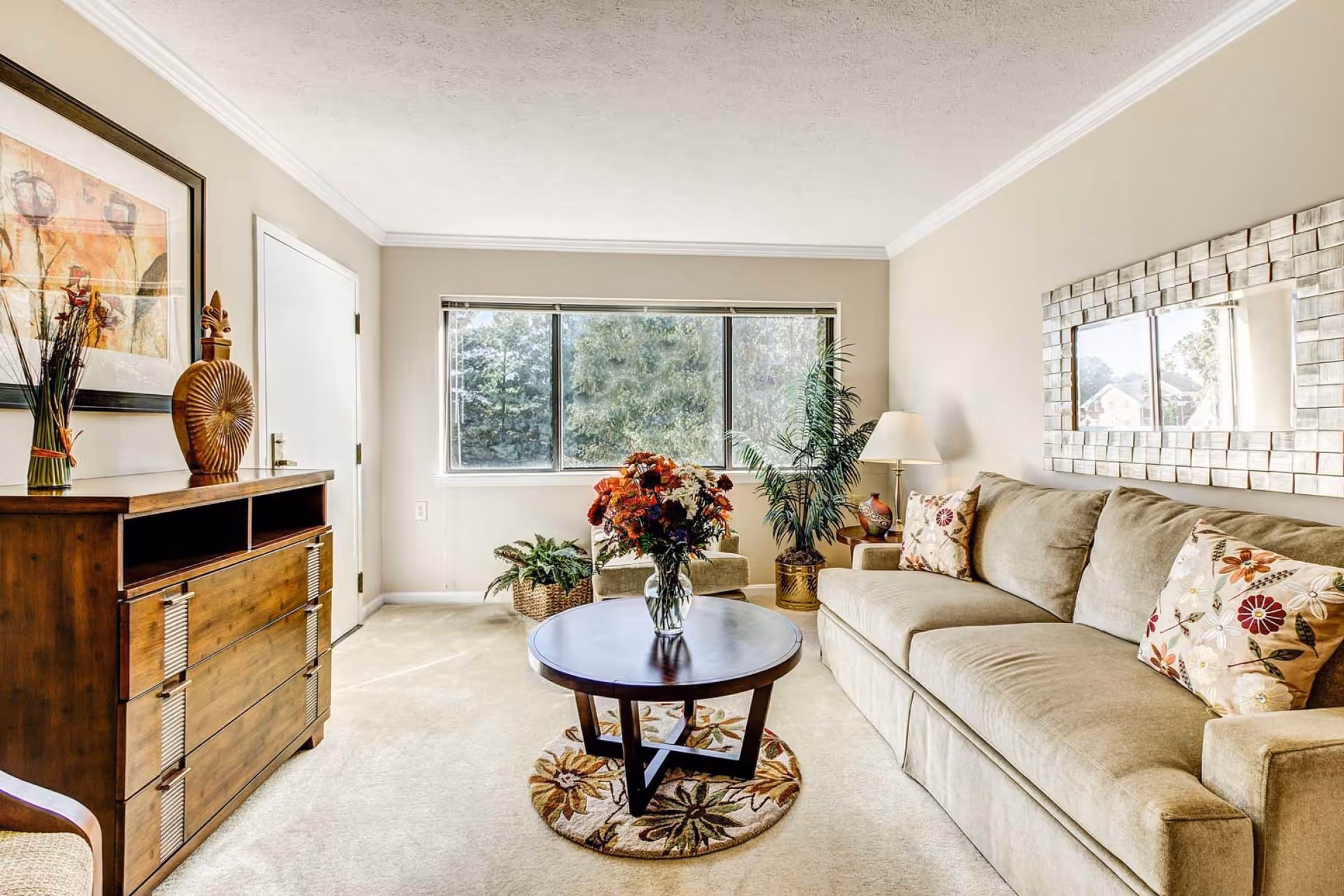 A bright living room with a beige sofa adorned with floral cushions, a round wooden coffee table with a vase of colorful flowers, a large window letting in natural light, a wooden dresser with decorative items, a floor lamp, and a large mirror on the wall.
