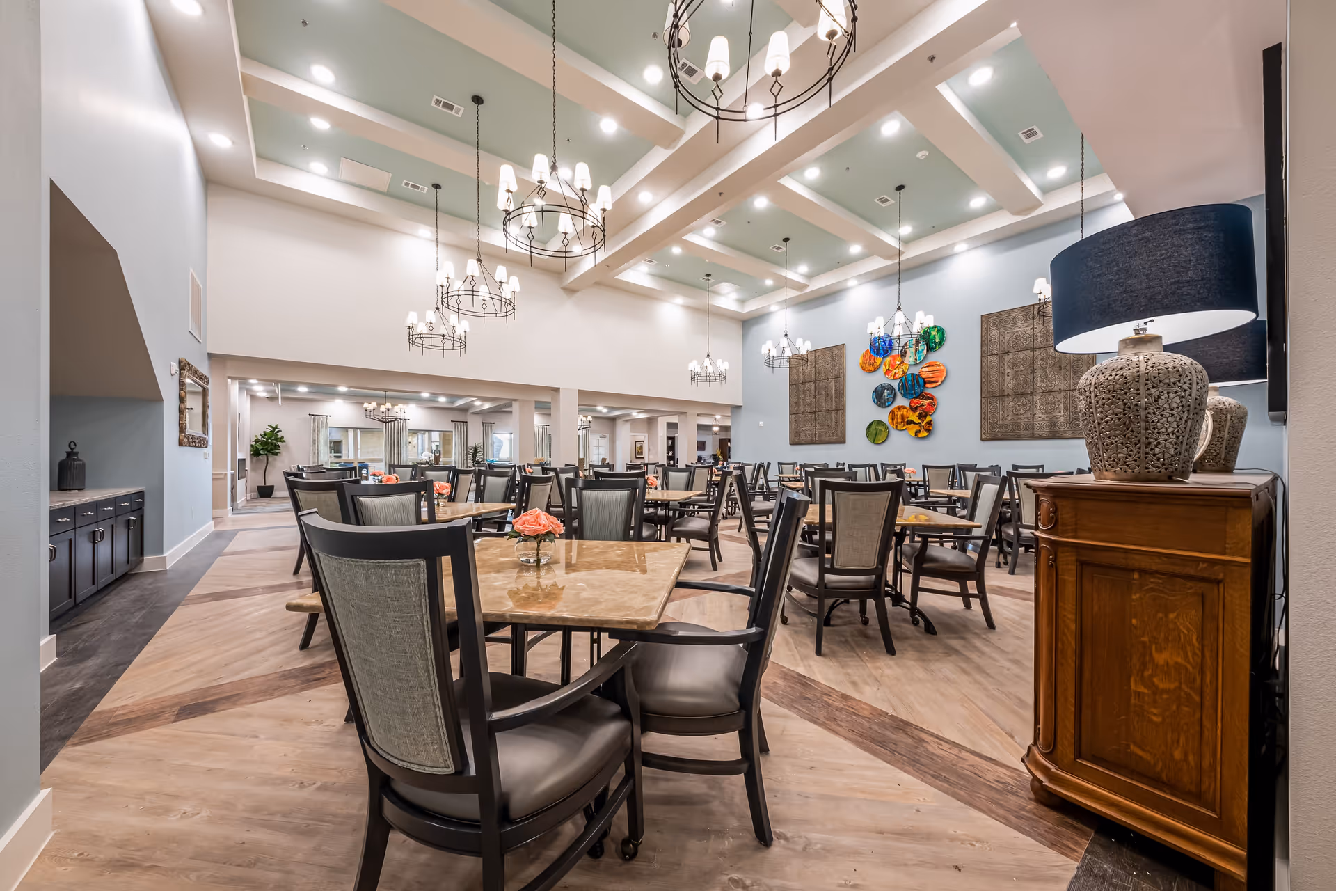A spacious, well-lit dining room with multiple tables and chairs arranged neatly. The ceiling features recessed lighting and decorative chandeliers. The walls are adorned with colorful circular art and large square panels. A wooden cabinet with a decorative lamp is visible on the right side.