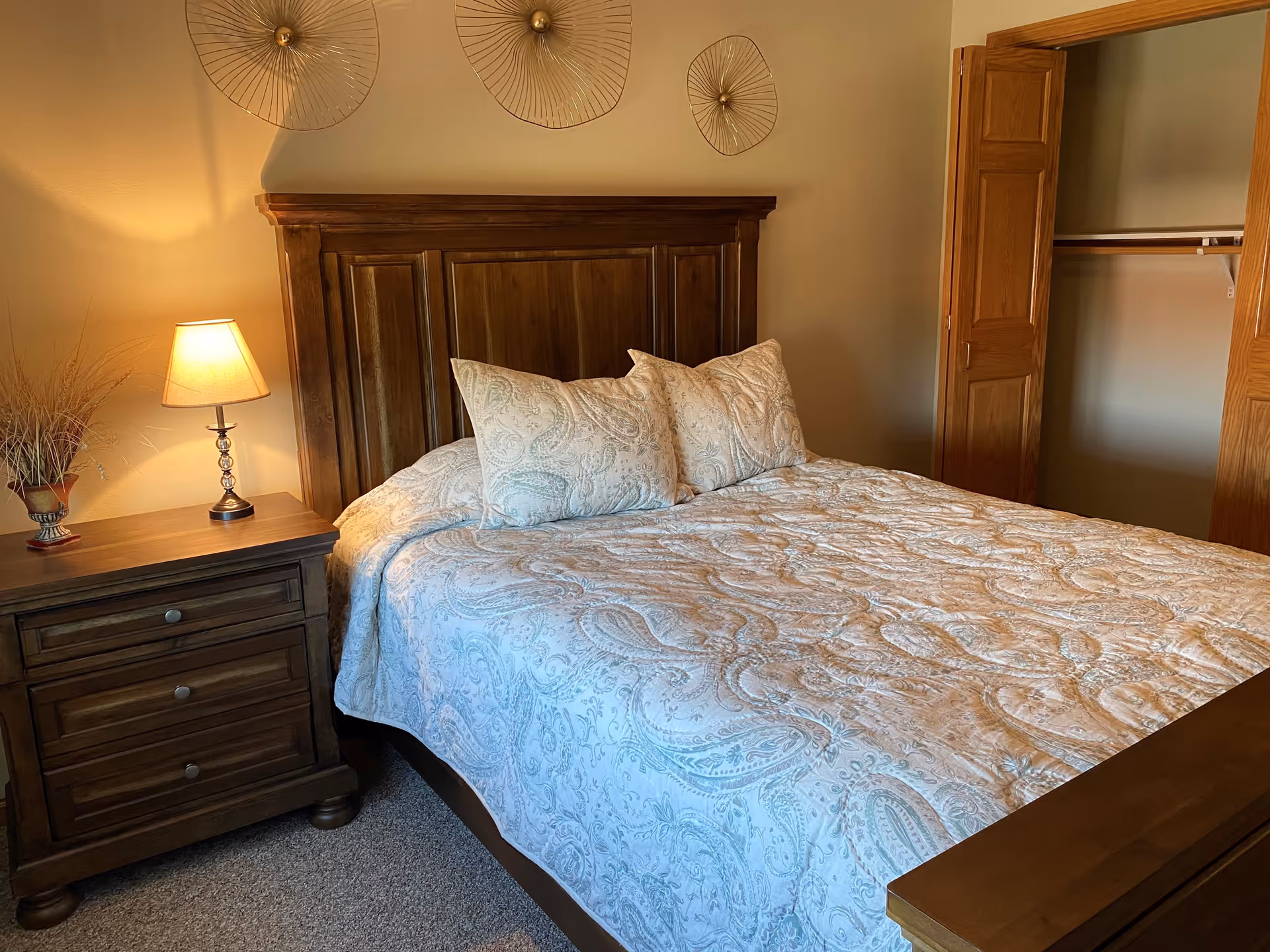 A cozy bedroom with a wooden bed frame and headboard, a bed covered with a light blue and white patterned quilt and matching pillows. To the left of the bed is a wooden nightstand with three drawers, a table lamp with a beige shade, and a decorative vase with dried plants. On the wall above the headboard are three circular metal wall decorations. To the right is an open wooden closet door revealing an empty closet with a hanging rod.
