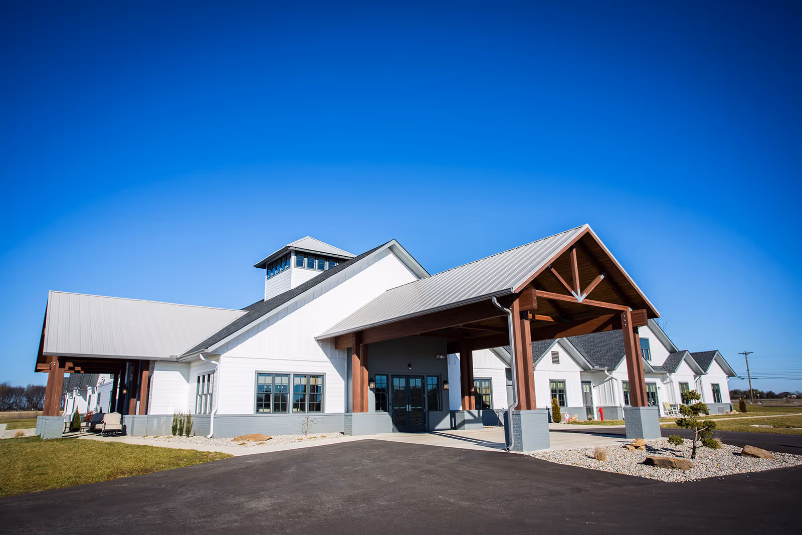 Exterior view of Lewis Manor Assisted Living facility showing a large white building with a gray metal roof and wooden beams supporting a covered entrance. The building is surrounded by a paved driveway, some landscaping with rocks and small plants, and a clear blue sky overhead.