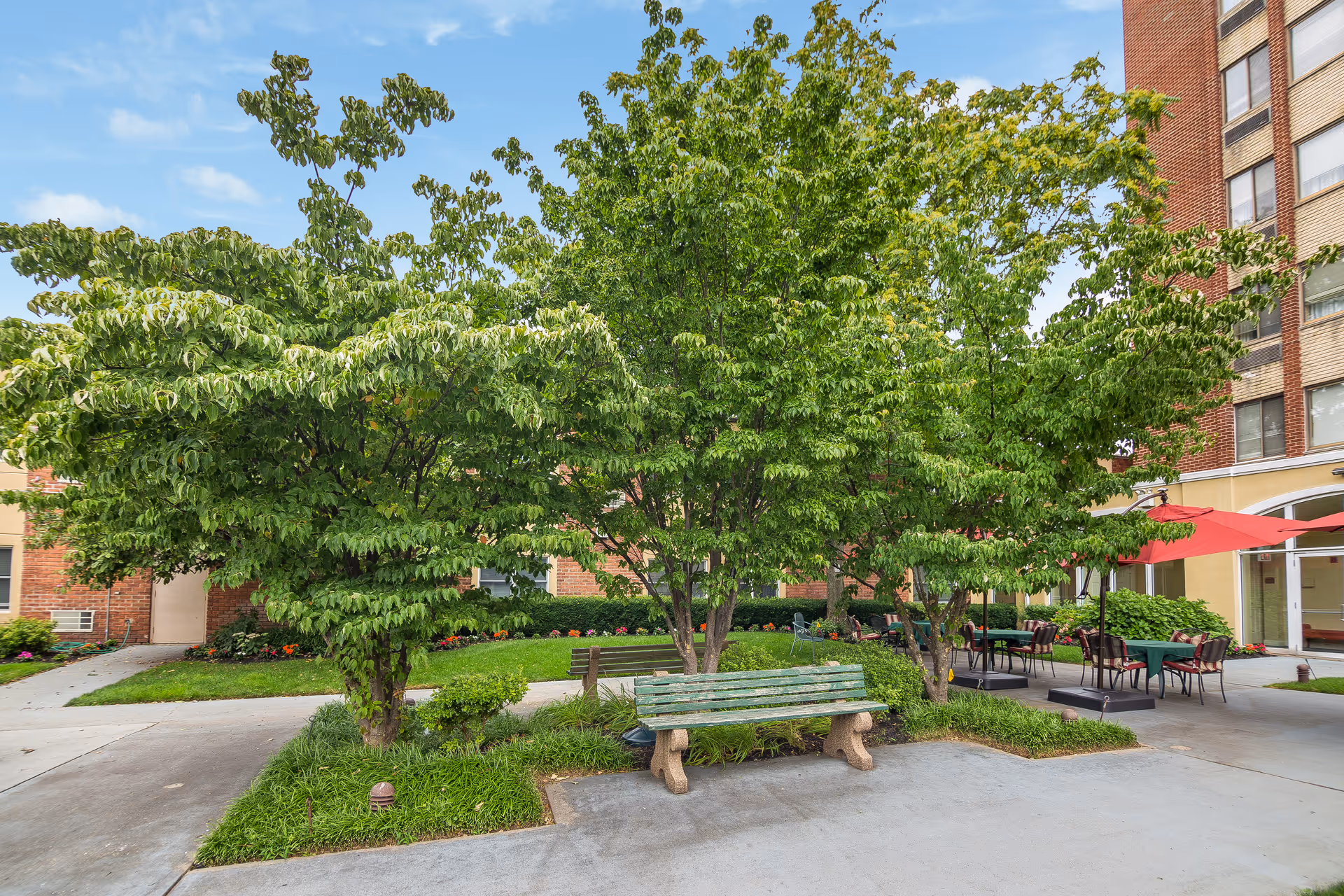 Outdoor courtyard area at Castle Senior Living featuring a green wooden bench surrounded by trees and shrubs, with a paved walkway and patio tables with red umbrellas near the building entrance.