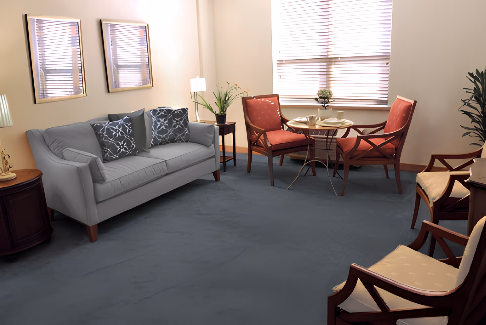 A cozy living room with a gray sofa adorned with patterned pillows, two red cushioned wooden chairs around a small round table set with cups and plates, two cream cushioned wooden chairs, a side table with a lamp and plant, and a window with blinds letting in natural light.