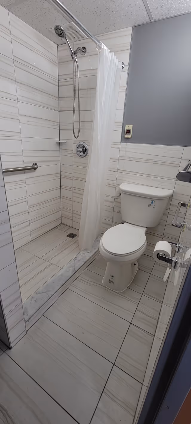 A clean bathroom with a walk-in shower featuring a handheld showerhead and a white shower curtain. The walls and floor are tiled with light-colored tiles with subtle horizontal stripes. There is a white toilet next to the shower, a toilet paper holder mounted on the wall, and a grab bar inside the shower for safety.
