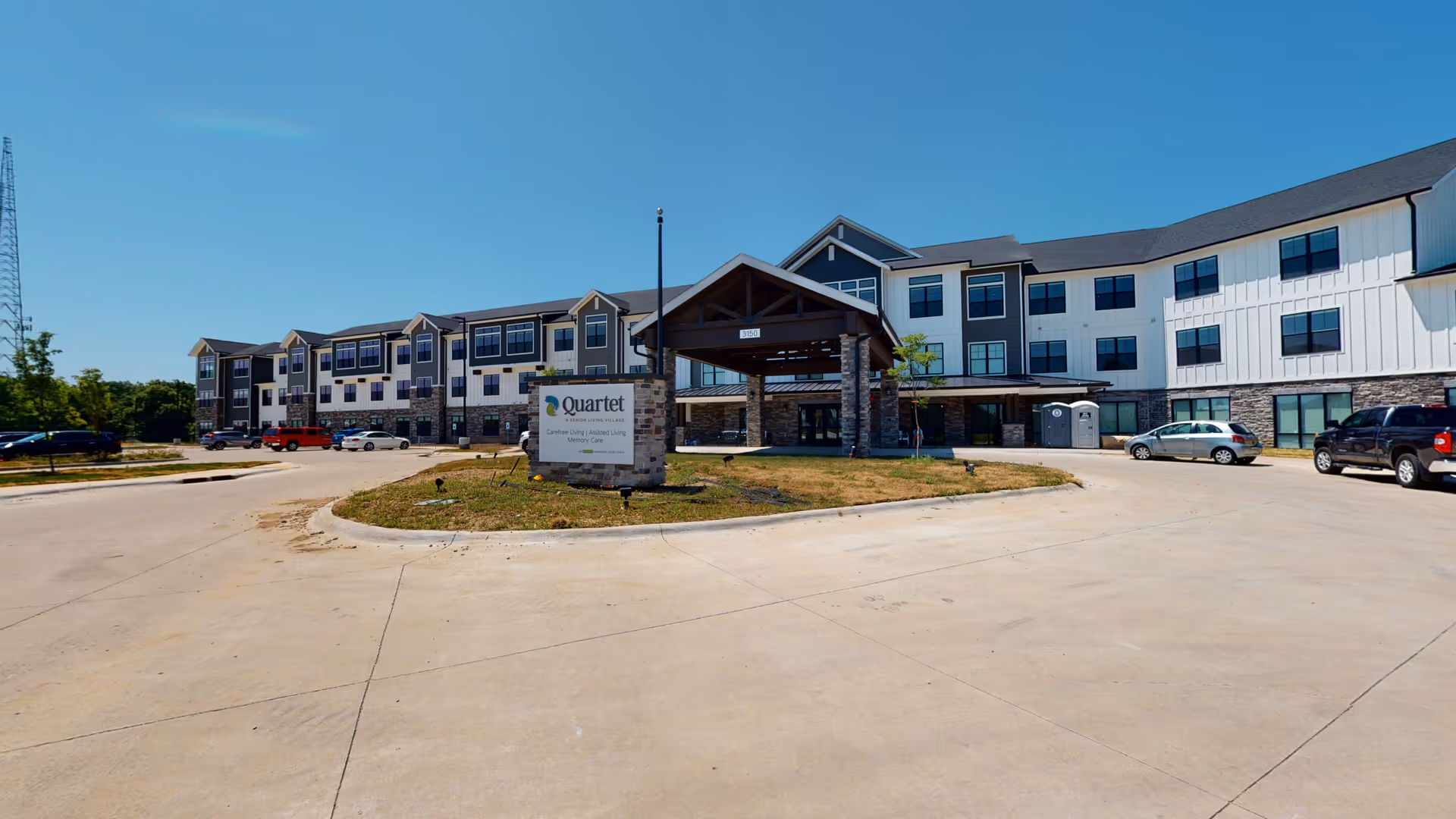 Exterior view of Quartet - A Senior Living Village building under a clear blue sky, showing a large three-story structure with multiple windows, a covered entrance supported by stone pillars, a parking area with several cars, and a sign in front displaying the facility's name.