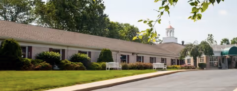 Exterior view of a single-story senior living facility building with a brown roof, white benches along the front, well-maintained green bushes and lawn, and a small cupola on the roof. The sky is clear with some tree branches visible in the foreground.