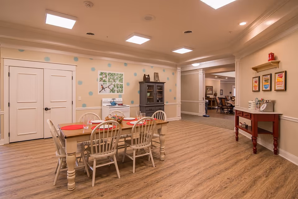 A dining area in a senior living facility with a wooden table set for six with red placemats and glasses. The room has light-colored walls decorated with blue polka dots and a picture of birds on a branch. There is a white stove and a dark gray cabinet against the wall. To the right, a red side table holds various items including a scale and a fire extinguisher on a small shelf above it. The floor is wood-patterned, and the ceiling has recessed lighting. An open doorway leads to another room with more seating visible.